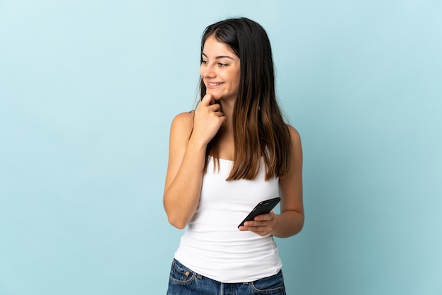 Young caucasian woman using mobile phone isolé sur bleu à la recherche sur le côté et souriant