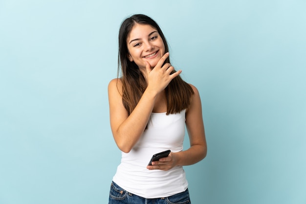 Young caucasian woman using mobile phone isolé sur fond bleu heureux et souriant