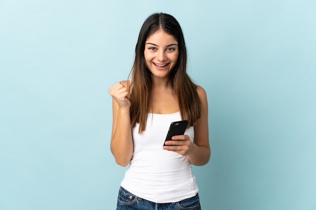 Young caucasian woman using mobile phone isolé sur mur bleu célébrant une victoire en position de gagnant