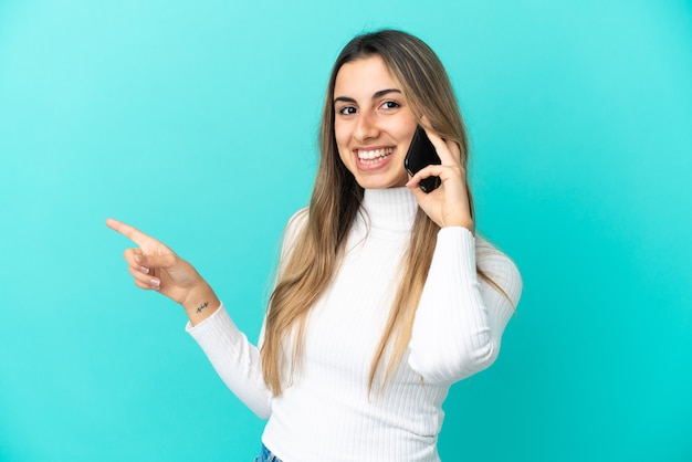 Young caucasian woman using mobile phone isolé sur un mur bleu pointant le doigt sur le côté