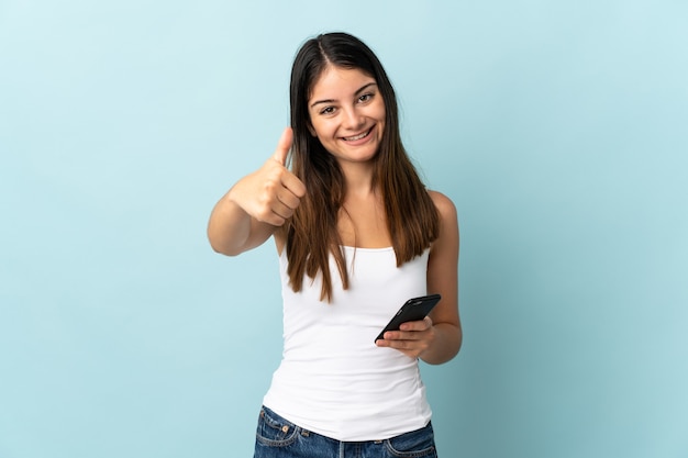 Young caucasian woman using mobile phone isolé sur mur bleu avec les pouces vers le haut parce que quelque chose de bien s'est passé