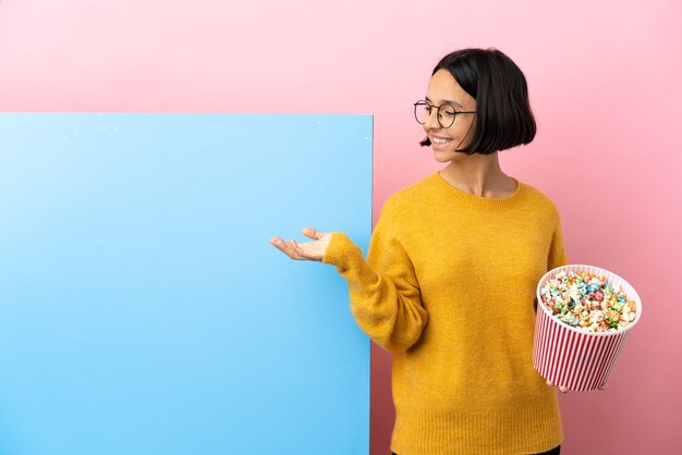 Young mixed race woman holding pop-corn avec une grande bannière sur mur isolé étendant les mains sur le côté pour inviter à venir