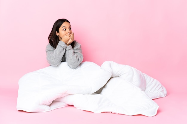 Young mixed race woman wearing pijama assis sur le plancher couvrant la bouche et regardant sur le côté