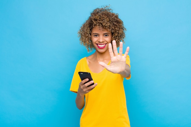 Young smiling and looking friendly, montrant le numéro cinq ou cinquième avec la main vers l'avant, compte à rebours avec un téléphone portable