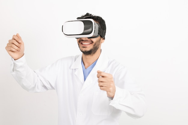 Young smiling male doctor in whitecoat et casque vr à la recherche d'informations sur l'affichage virtuel lors de la présentation