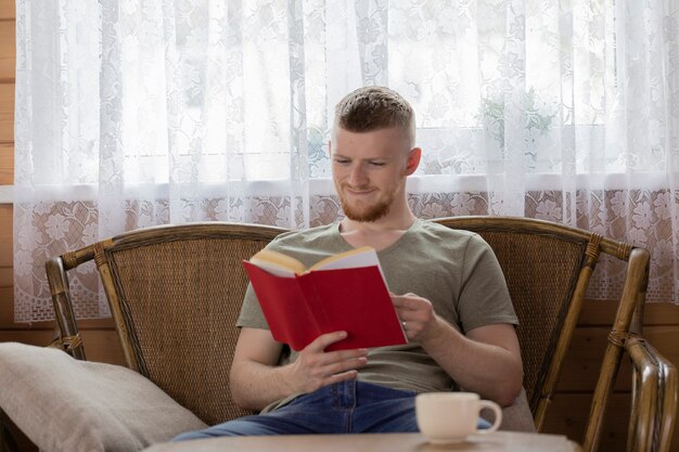 Young smiling man reading book avec couverture rouge sur banc en osier dans la campagne maison en bois