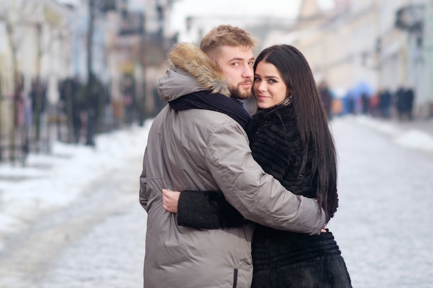 Young teen couple hug eachother standing in center of street uner red umbrella in winter under snowfall, regardant dans les yeux les uns aux autres et souriant