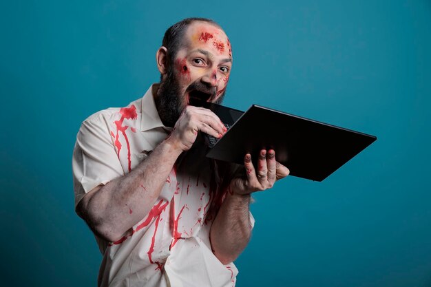 Zombie effrayant mordant sur un ordinateur portable, ayant des cicatrices sanglantes et des blessures sur un visage sale et effrayant. Monstre mort-vivant effrayant étant agressif avec le pc, semblant dangereux, cruel et mortel.