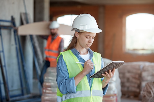 a construction industry maintenance engineer is a pretty woman dressed in a uniform and protective helmet with a tablet in her hands against the  of a construction site and a worker