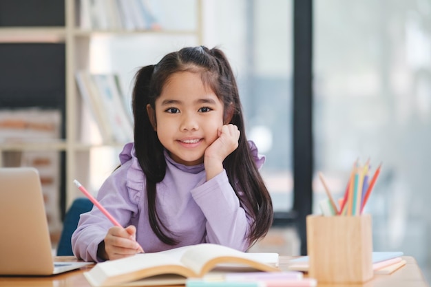 A young girl is sitting at a desk with a laptop and a book