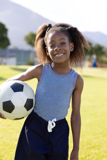 African American child holding soccer ball on grassy sports field smiling under clear sky