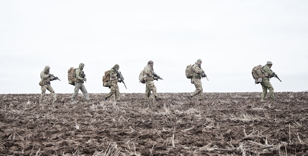Army soldiers on march. Elite forces fighters group, commando tactical unit, reconnaissance team members in camouflage uniform, walking in line, carrying backpacks on muddy terrain