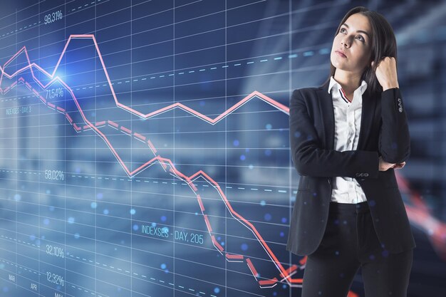 Attractive thoughtful young european businesswoman looking at glowing downward red forex chart on blurry office interior background Crisis and stock market fall concept Double exposure