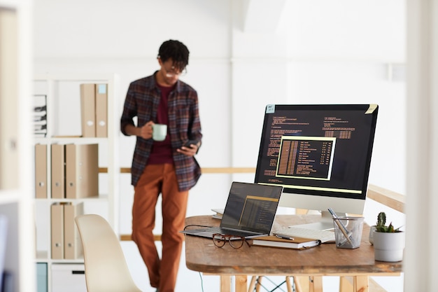 Background image of programming code on computer screen in modern office interior with blurred shape of African-American man, copy space