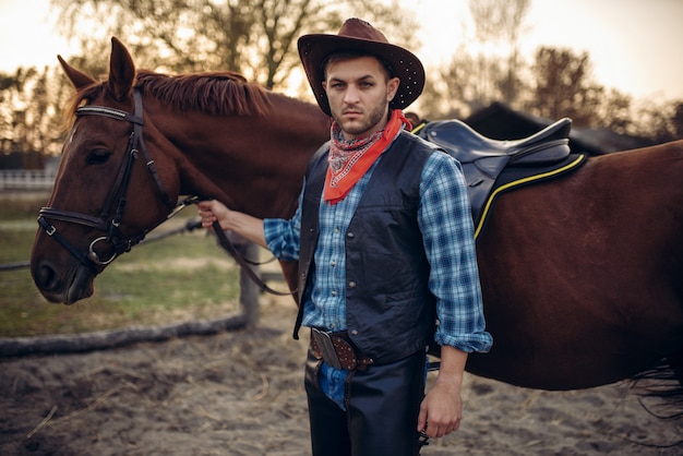 Brutal cowboy in jeans and leather jacket poses with horse on texas ranch, western. Vintage male person with animal, wild west