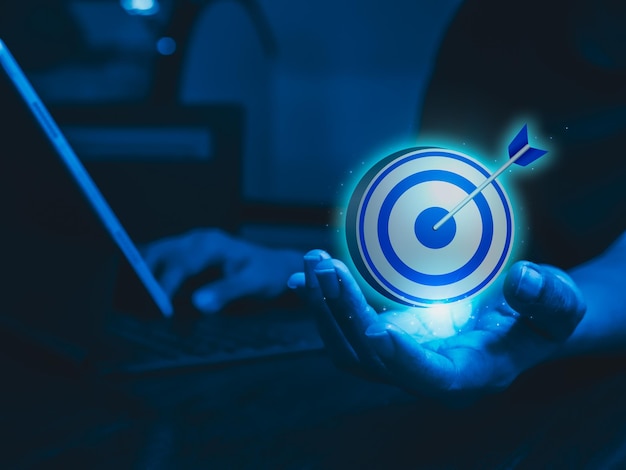 Businessman hand holding big goal target dart icon while working with laptop computer blue tone Business strategy action plan and future innovation technology with goal and success concepts