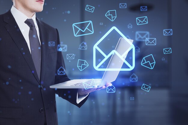 Businessman hands using laptop with glowing email letter icons on blurry office interior background Communication and business message concept