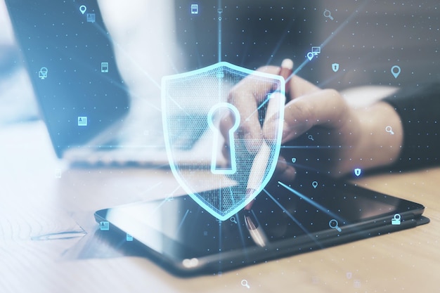 close up of male hand at wooden office desktop using laptop and tablet with creative glowing web security shield on blurry background secure information concept double exposure