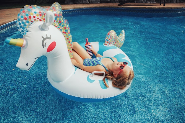 Cute adorable girl in sunglasses with drink lying on inflatable ring unicorn kid child enjoying