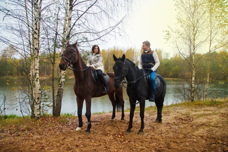 Foto cute young couple on horsebacks in the autumn forest by lake. riders in autumn park in inclement cloudy weather with light rain. concept of outdoor riding, sports and recreation. copy space