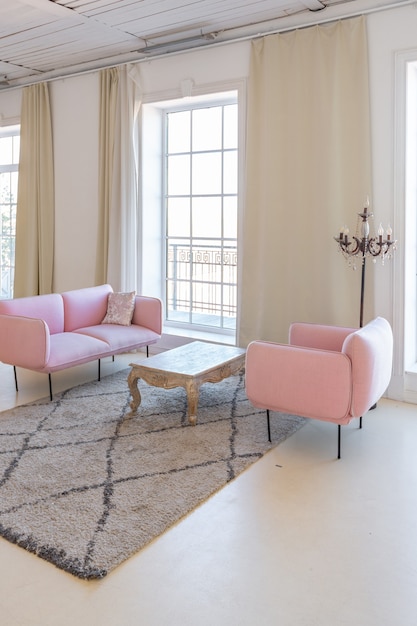 Delicate and cozy light interior of the living room with modern stylish furniture of pastel pink color and white walls with stucco moldings in daylight