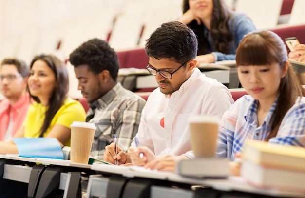 education, high school, university, learning and people concept - group of international students with notebooks and coffee writing test in lecture hall