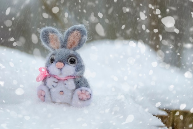 funny rabbit in snow, christmas or easter concept. a toy gray hare sits in a snowdrift against the backdrop of a forest and falling snow. postcard printable with place for text.