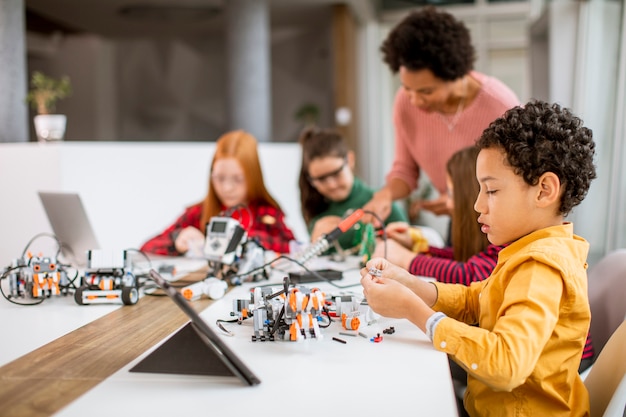Group of happy kids with their African American female science teacher with laptop programming electric toys and robots at robotics classroom