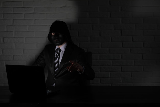 Hacker in black mask and hood at the table in front of the monitor