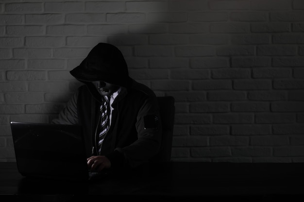 Hacker in black mask and hood at the table in front of the monitor