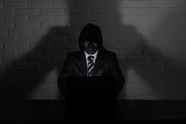 Hacker in black mask and hood at the table in front of the monitor