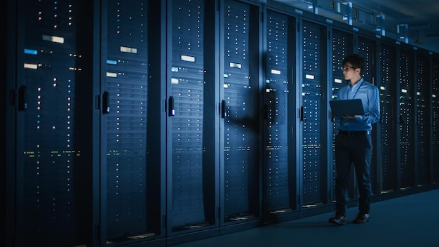 In Dark Data Center Male IT Specialist Walks along the Row of Operational Server Racks Uses Laptop for Maintenance Concept for Cloud Computing Artificial Intelligence Supercomputer Cybersecurity