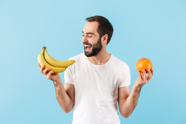 Kjekk og blid ung skjeggete mann iført t-skjorte som står isolert over en blå vegg, viser appelsin og banan