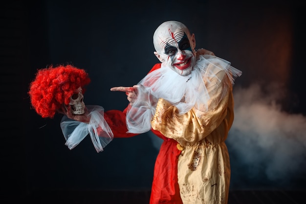 mad bloody clown holds human skull in red wig, horror. man with makeup in carnival costume, crazy maniac
