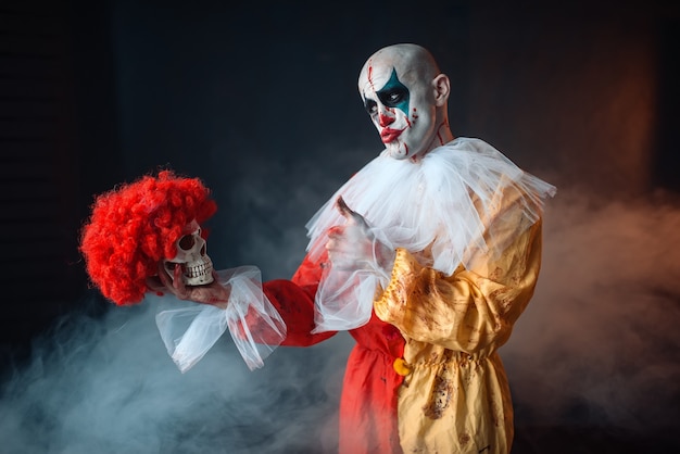 mad bloody clown holds human skull in red wig