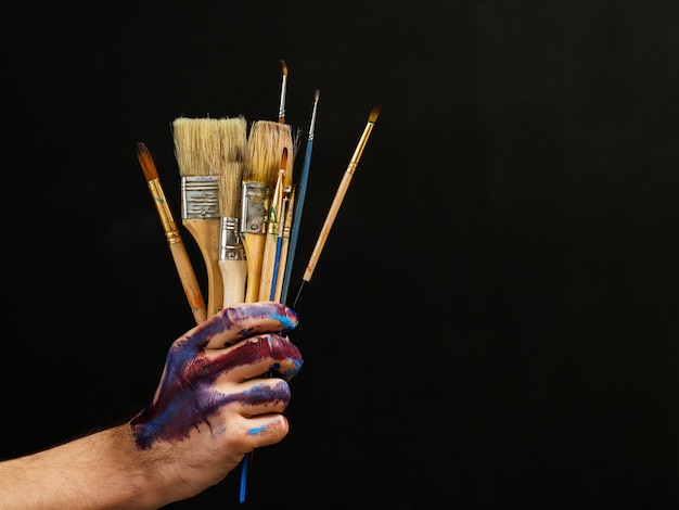 Modern art. Painter essential tools. Closeup of bunch of paintbrushes in male hand over dark background.