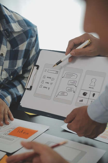 Nærbilde av UX-utvikler og UI-designer som brainstormer om wireframe-design for mobilappgrensesnitt på bord med kundebrief og fargekode på moderne kontorKreativt digitalt utviklingsbyrå