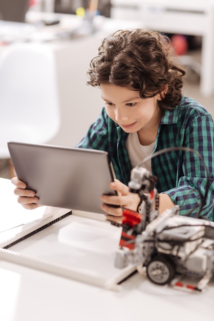 Searching for creative ideas. Smiling cheerful concentrated boy sitting in the classroom and using gadget while studying and expressing joy