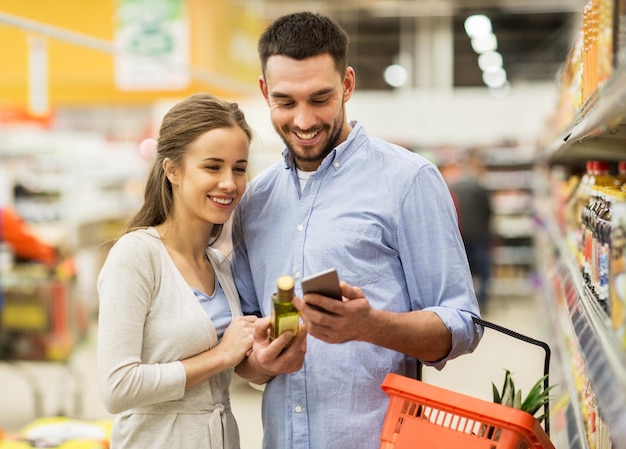 shopping, food, sale, consumerism and people concept - happy couple with smartphone buying olive oil at grocery store or supermarket