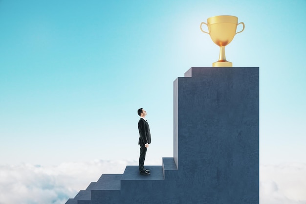 Side view of young european man standing on stairs to golden winner's cup with obstacle on blue sky background Success and career growth concept