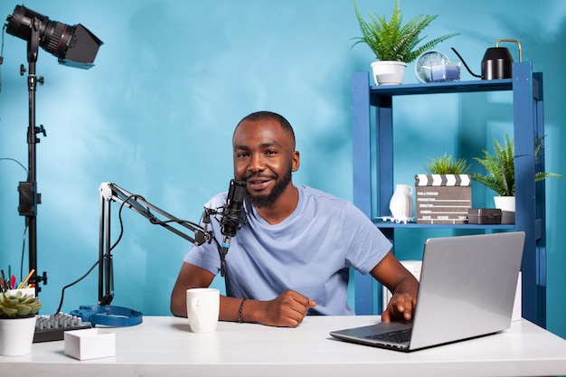 Smiling influencer interacting with audience during online live show looking at camera with one hand using laptop sitting at desk with broadcasting setup. Content creator talking with fanbase in recor