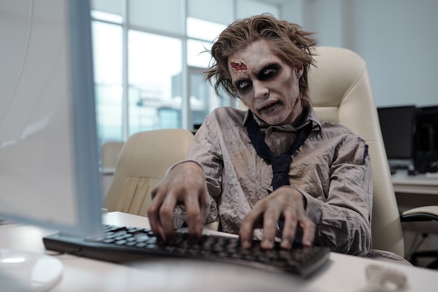 Spooky businessman with zombie body painting looking at computer screen and pressing keyboard buttons in office environment