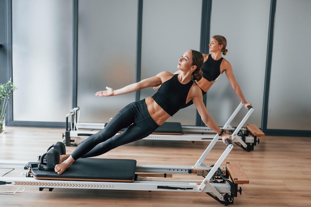 Standing on gym equipment and doing stretches Two women in sportive wear and with slim bodies have fitness yoga day indoors together