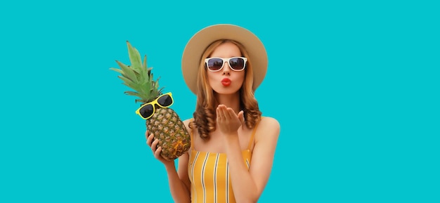 Summer portrait of beautiful young woman blowing a kiss posing with pineapple wearing hat glasses
