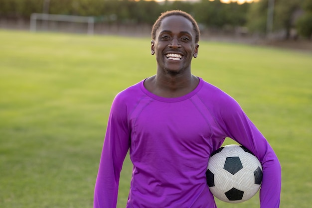 Teenage boy smiling and holding soccer ball on grassy soccer field under bright sky