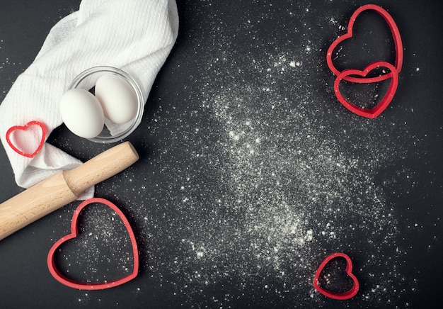 Valentine's Day cooking composition on the dark background with scattered flour. Flat lay, top view.