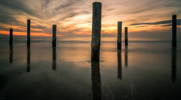 wooden posts amidst sea at beach against cloudy sky during sunset