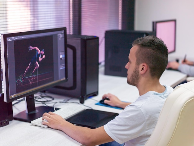 young graphic designer working at workplace. guy sitting at table retouching photo using digital tablet with pen and desktop computer indoors. freelancing and distance job concept