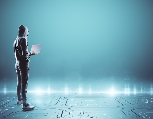 Young hacker with laptop in hoodie standing in abstract circuit interior with empty blue wall Copy space Hacking concept