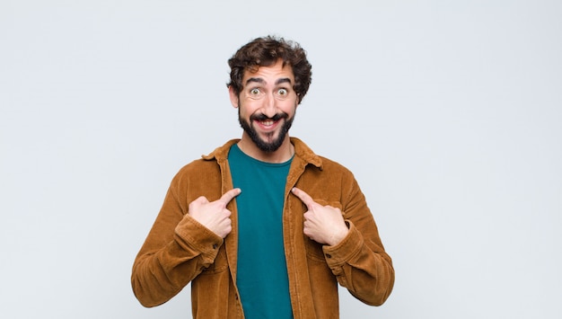 young handsome man feeling happy, surprised and proud, pointing to self with an excited, amazed look against flat wall